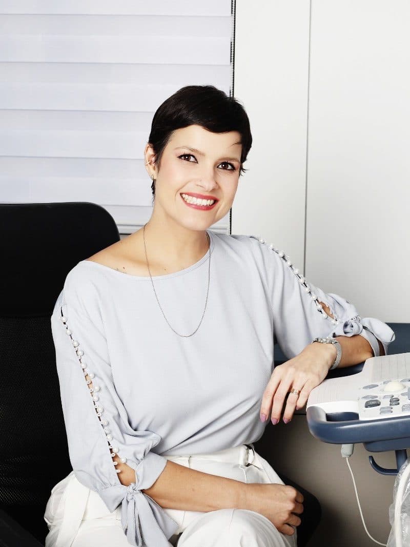 Dra. Michelle Sanches sorrindo no consultório, sentada ao lado do aparelho de ecocardiograma