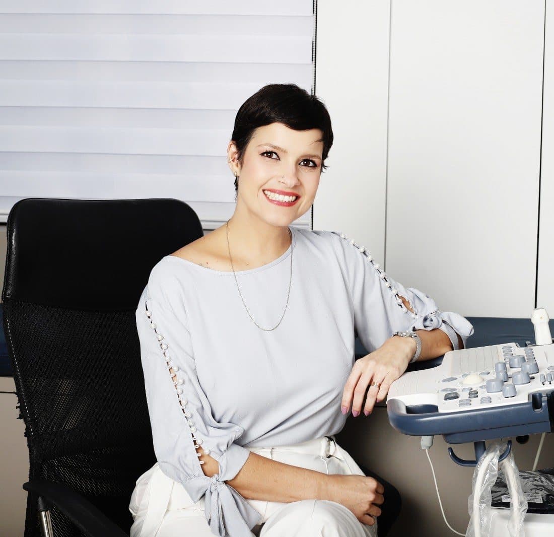 Dra. Michelle Sanches no consultório, perto da janela, ao lado do aparelho de ecocardiograma